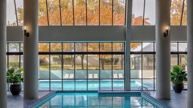 An indoor swimming pool with clear glass walls, potted plants, and a view of outdoor trees. - Marriott Albuquerque (Albuquerque)