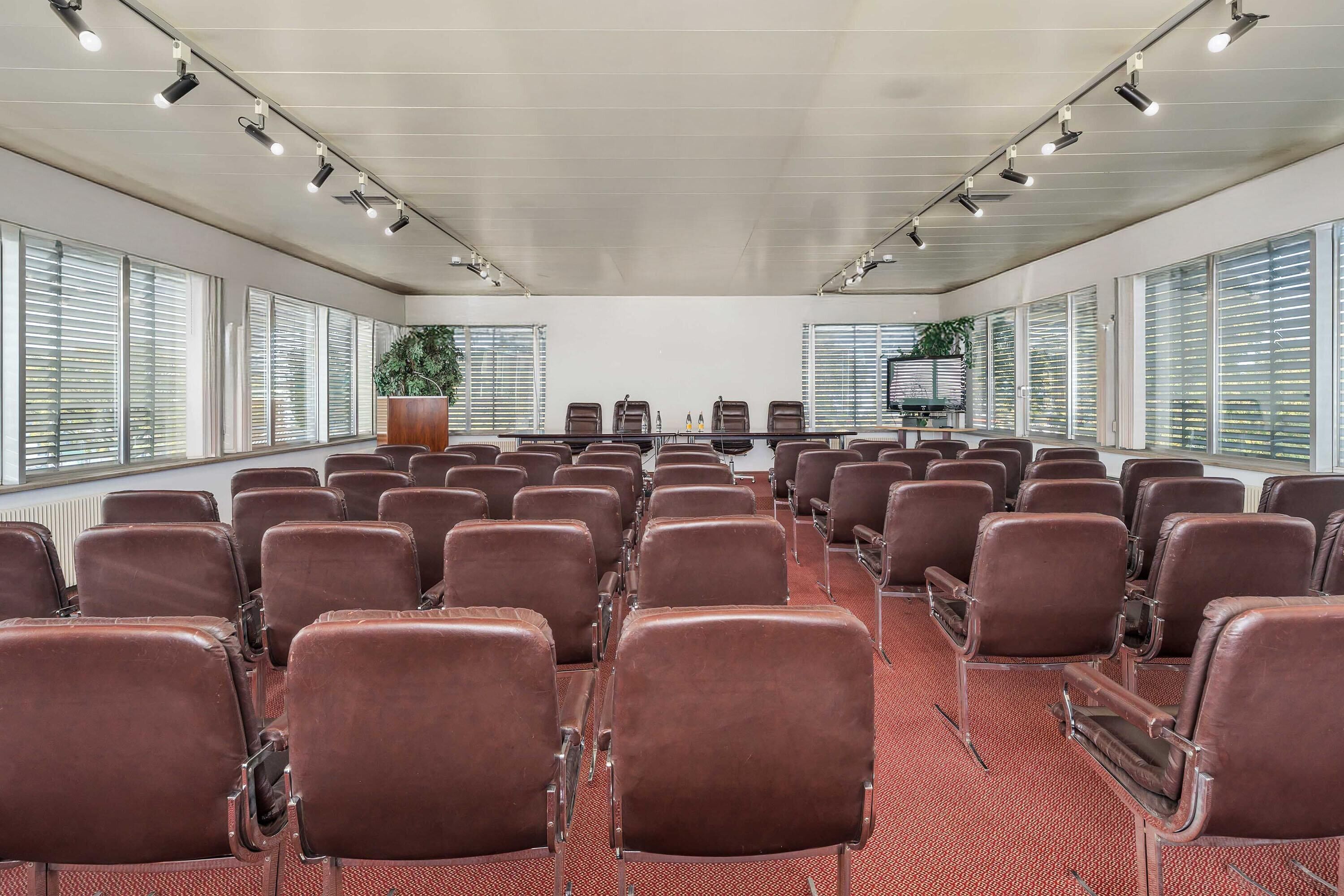 Een congreszaal met rijen bruine leren stoelen, een lange tafel en grote ramen met zonwering.
