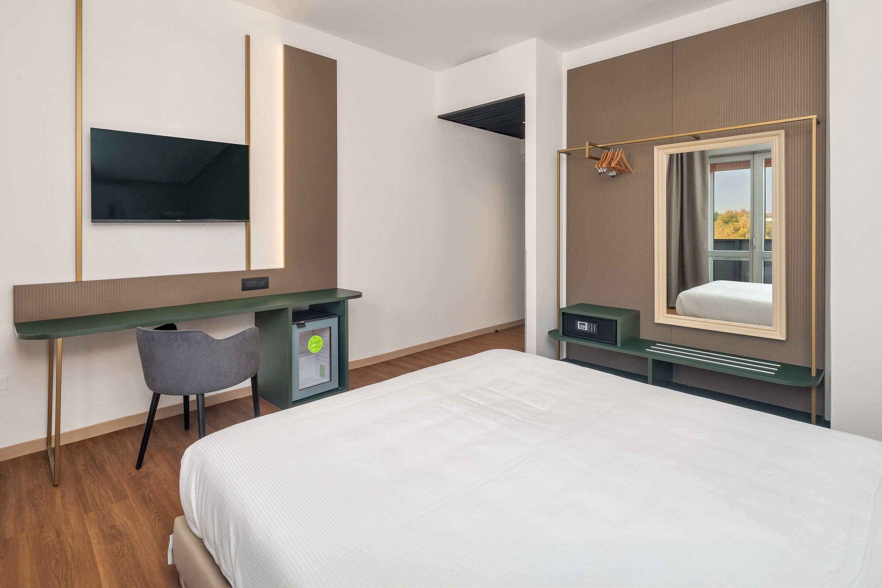 Ein modernes Hotelzimmer mit einem großen Bett, einem Schreibtisch mit Stuhl, einem Flachbildfernseher an der Wand und einem Fenster mit Blick ins Freie.