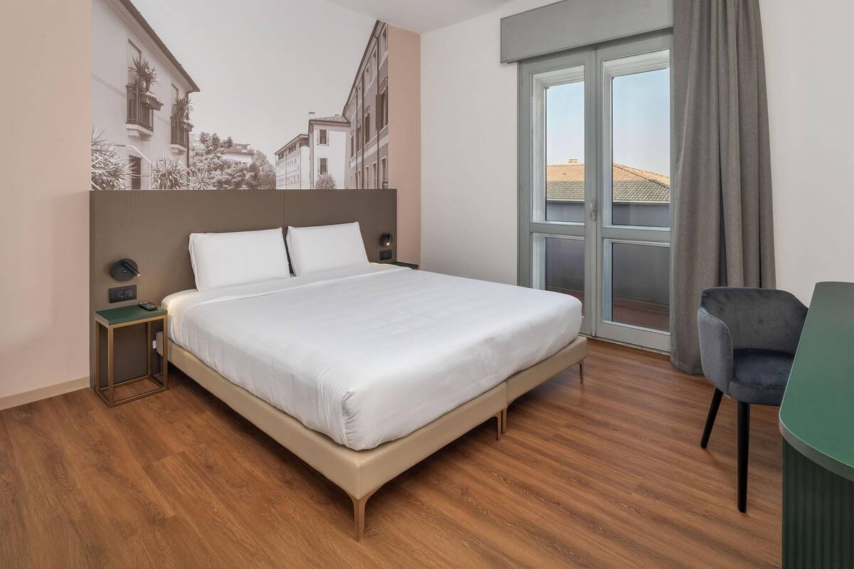 Ein modernes Hotelzimmer mit einem großen Bett, einem kleinen Schreibtisch und Blick auf umliegende Gebäude.