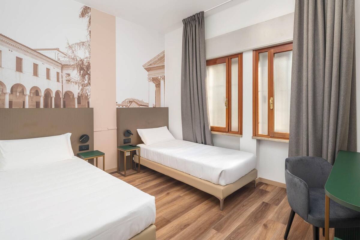 Ein Hotelzimmer mit zwei Einzelbetten, einem kleinen Tisch, einem Stuhl und einem Fenster mit Vorhängen.