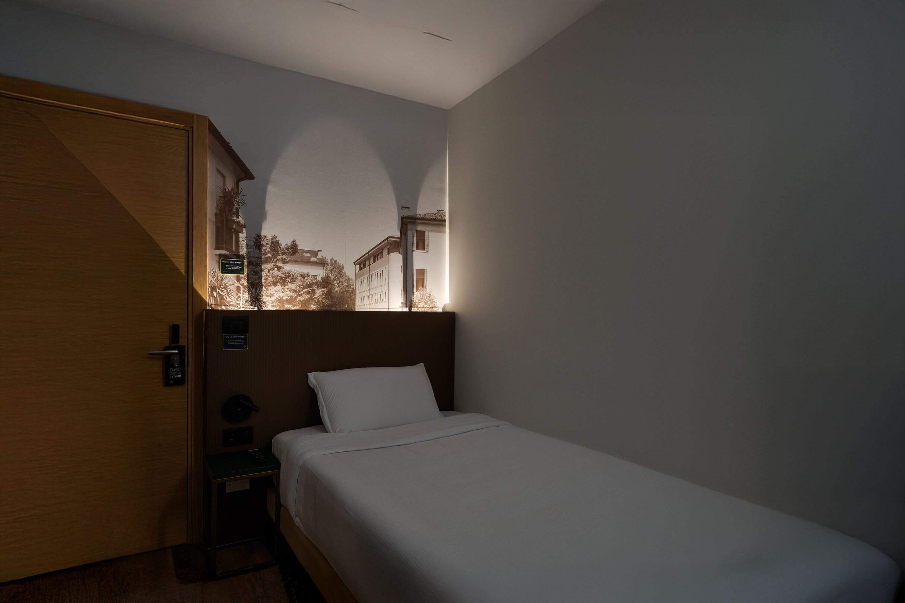 Une chambre d’hôtel avec un lit, une table de chevet, une porte en bois et une vue sur les immeubles par la fenêtre.