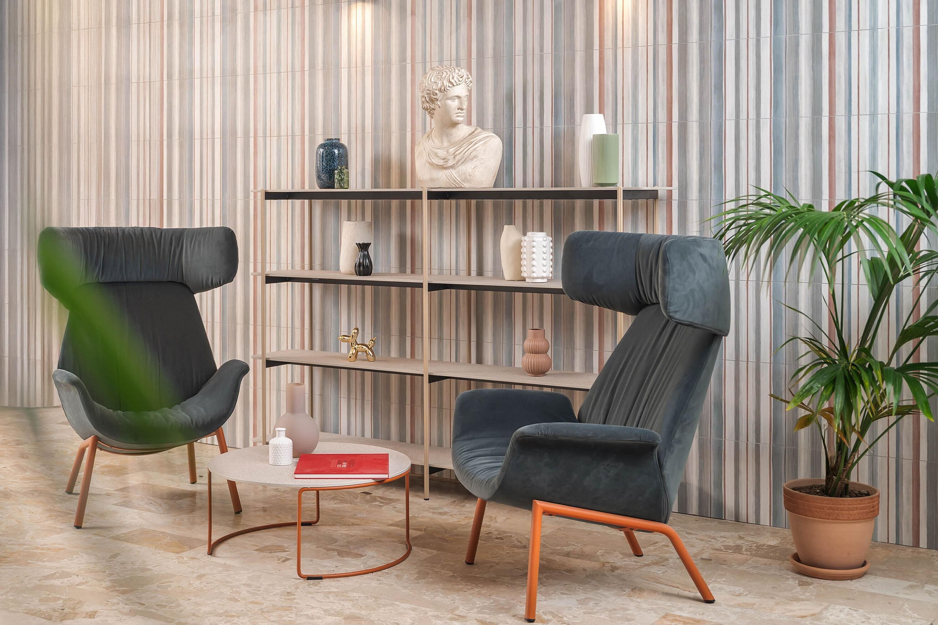 A modern room with two grey armchairs, a small round table, a bookshelf with decorative items, and a potted plant.