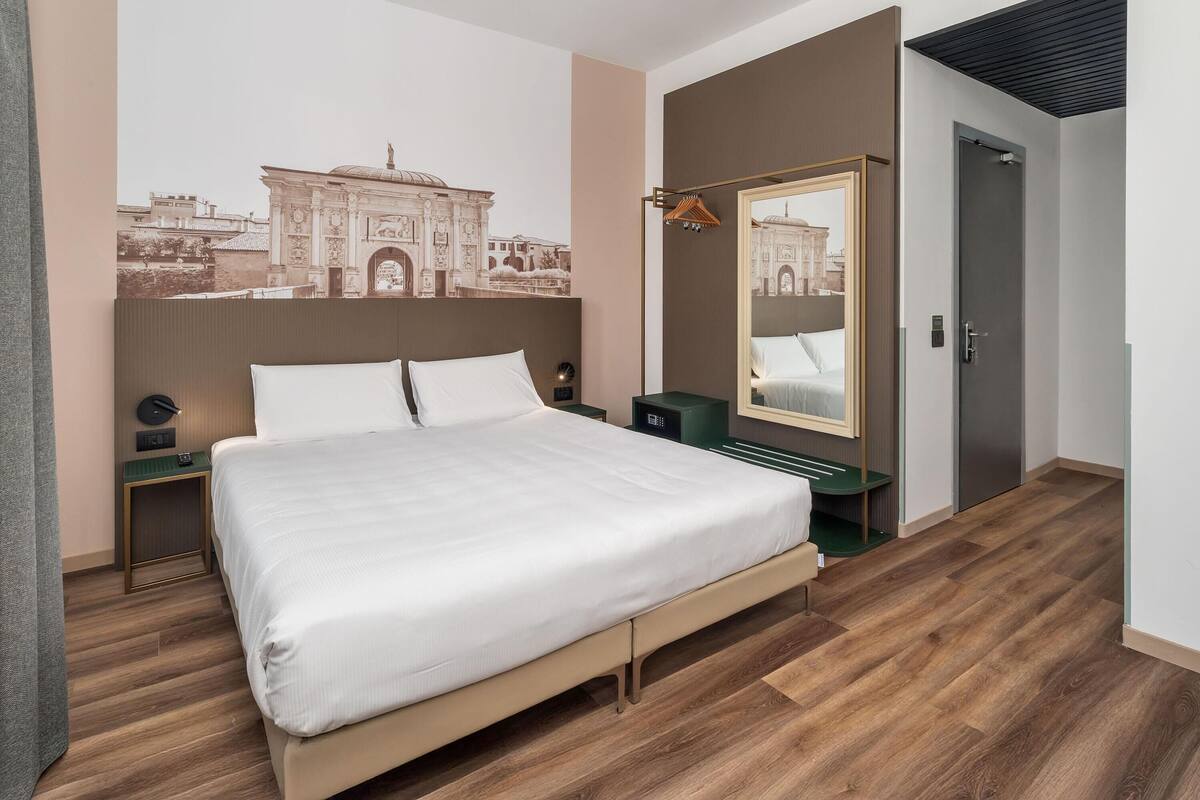 Ein modernes Hotelzimmer mit einem großen Bett, einem Nachttisch, einem Spiegel und einem Wandbild eines historischen Gebäudes.