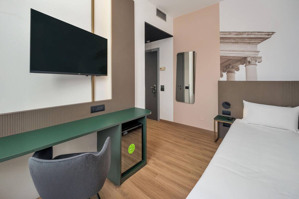 Ein Hotelzimmer mit Bett, Schreibtisch mit Stuhl, Wandfernseher, Spiegel und Nachttisch mit Lampe.
