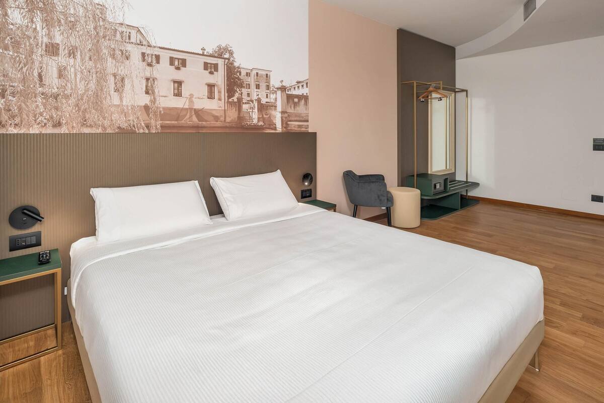Ein modernes Hotelzimmer mit einem großen Bett, einem Schreibtisch, einem Sessel, einem Spiegel und einem Wandbild.