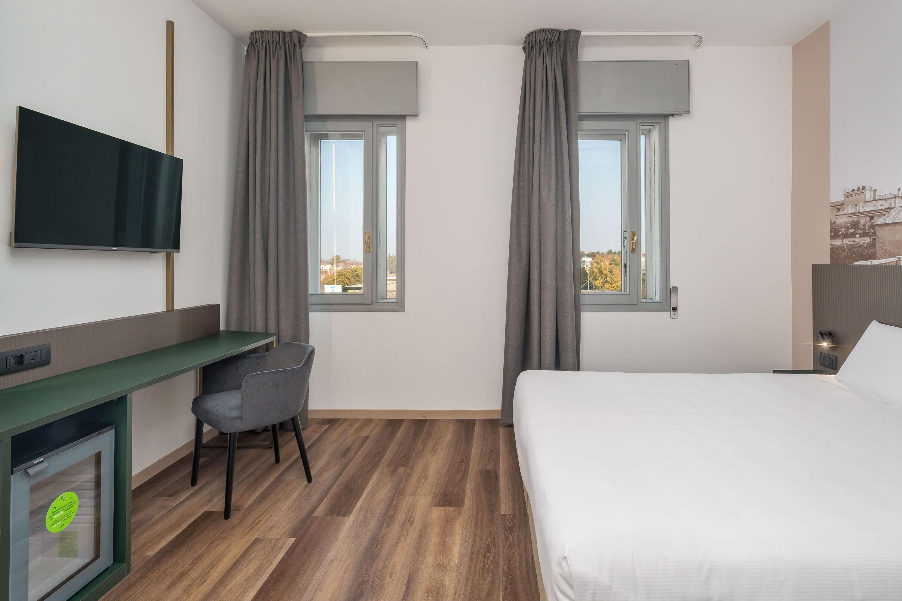 Ein Hotelzimmer mit Bett, Schreibtisch mit Stuhl, Wandfernseher und zwei Vorhängen versehene Fenster.