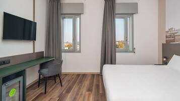 Ein Hotelzimmer mit Bett, Schreibtisch mit Stuhl, Wandfernseher und zwei Vorhängen versehene Fenster.