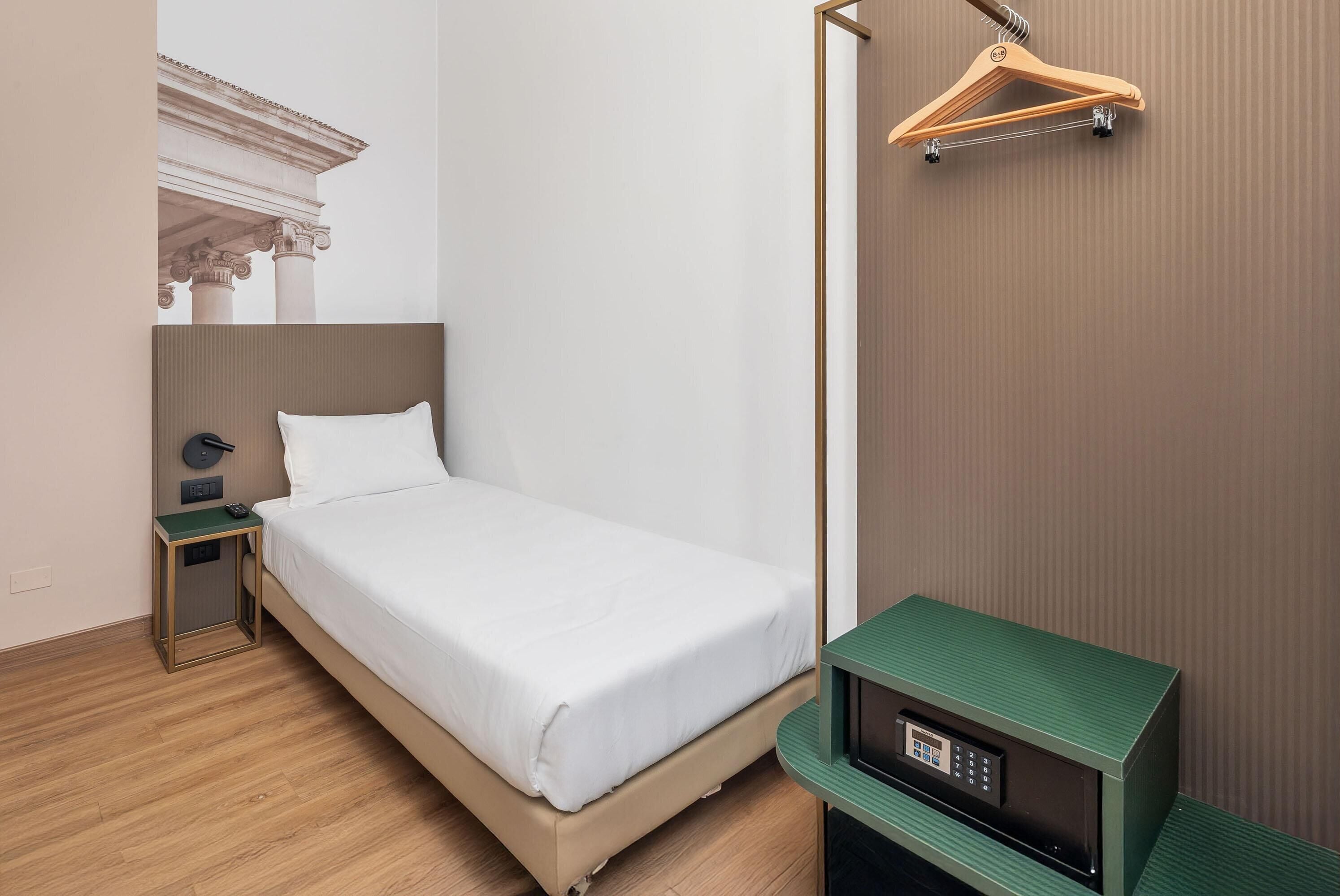 A hotel room with a bed, a nightstand, a green side table, a hanging wooden hanger, and a wall-mounted TV.