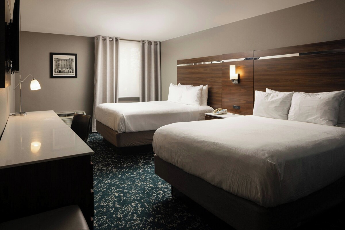 A hotel room with two beds, a desk, a lamp, a picture on the wall, and a window with curtains.