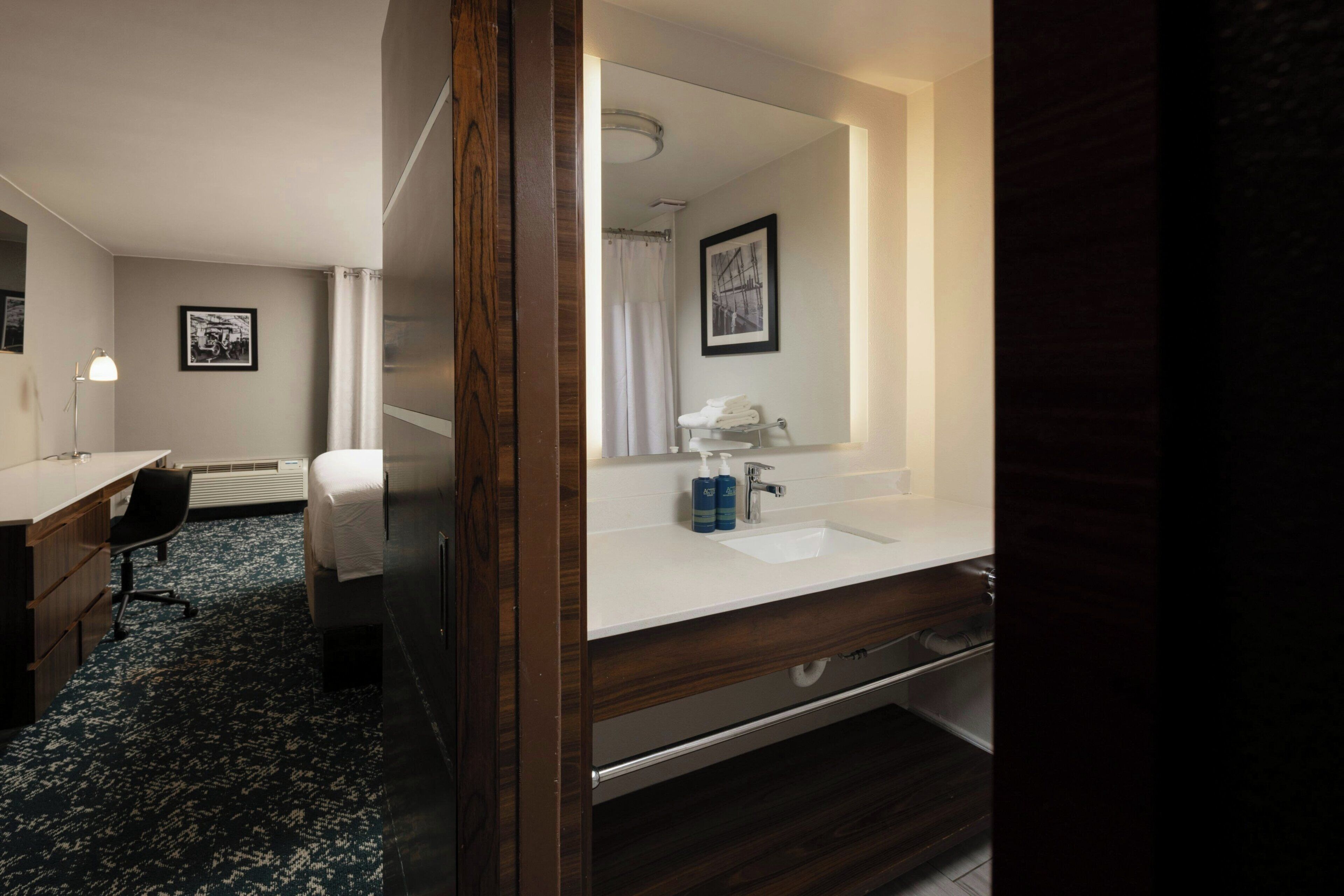 A hotel room with a bed, a desk, a chair, a bathroom with a sink and mirror, and a shower.