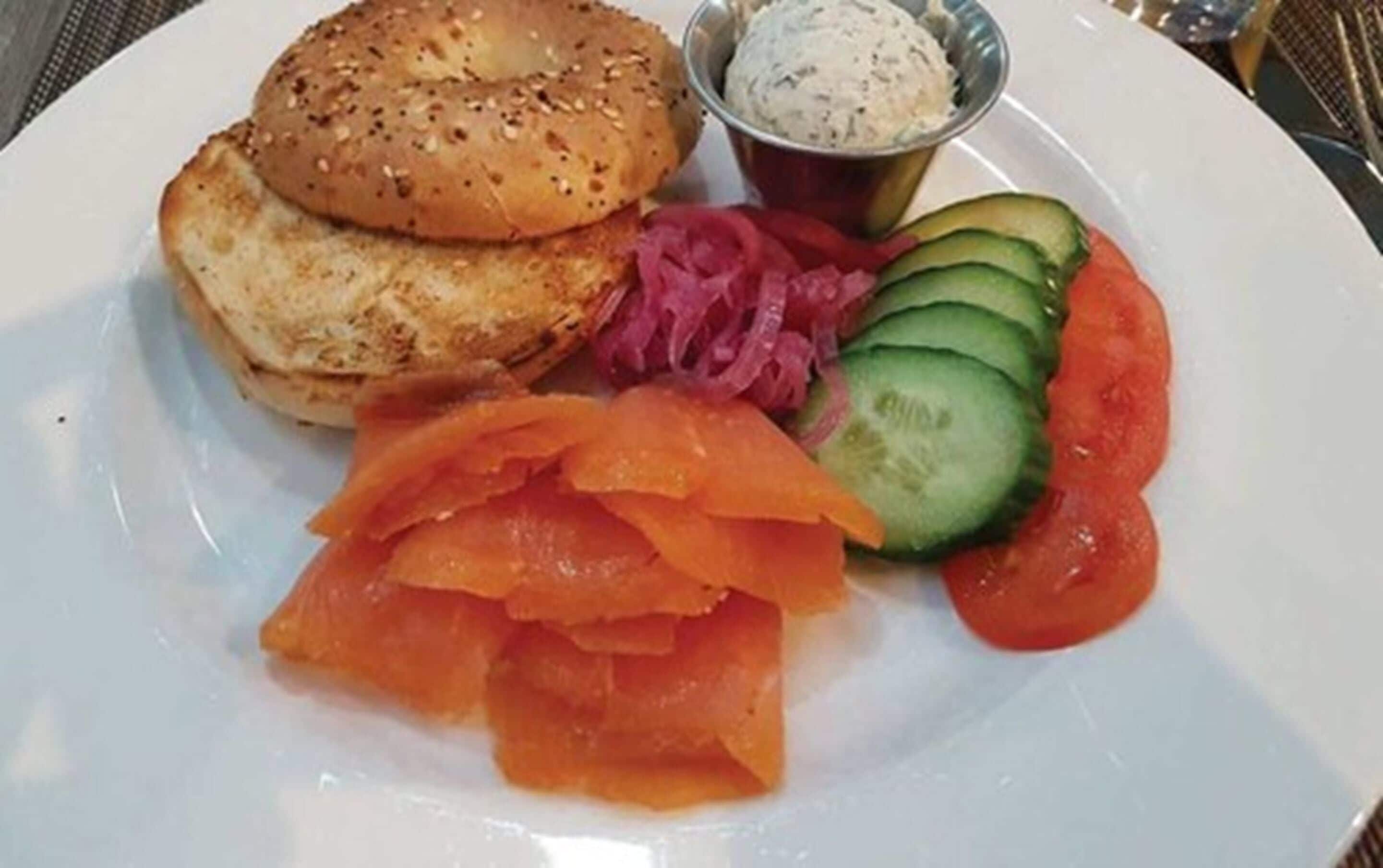 A plate with bagels, smoked salmon, cucumber slices, tomato, and a small bowl of cream cheese.