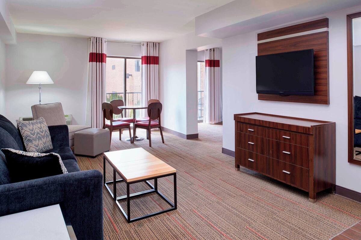 Habitación de hotel con sofá, una mesita, televisor montado en la pared y un mueble de madera.