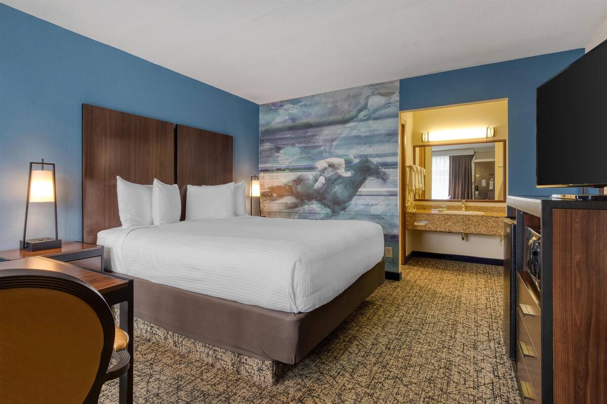 A hotel room with a large bed, a desk, a chair, a television, a mirror, and a bathroom with a sink and mirror.