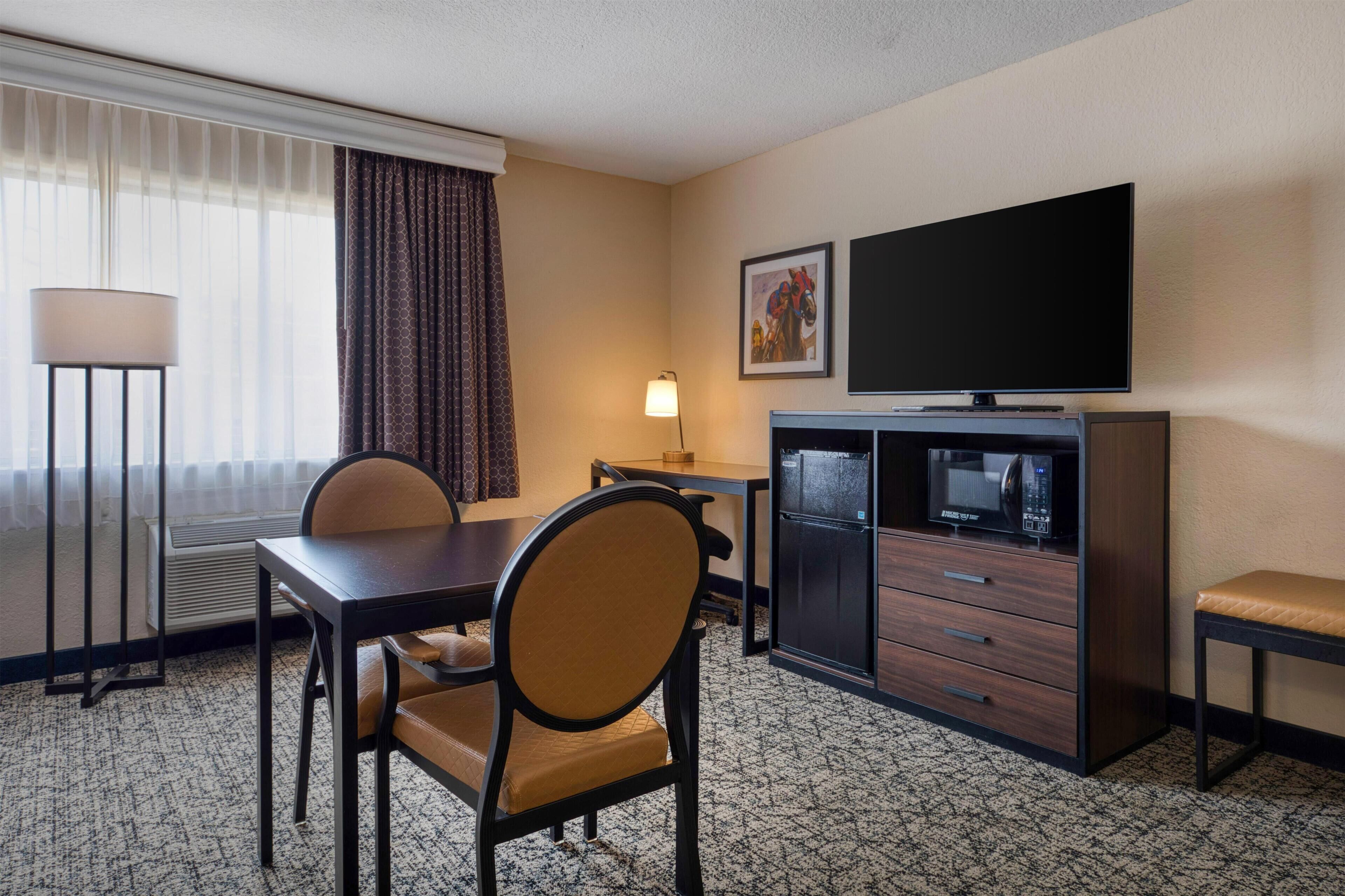 A hotel room with a dining table, chairs, a TV, a microwave, and a desk.