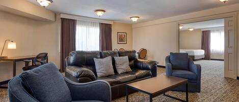 A hotel room with a leather sofa, a blue armchair, a small table, a desk with a lamp, and a large mirror.