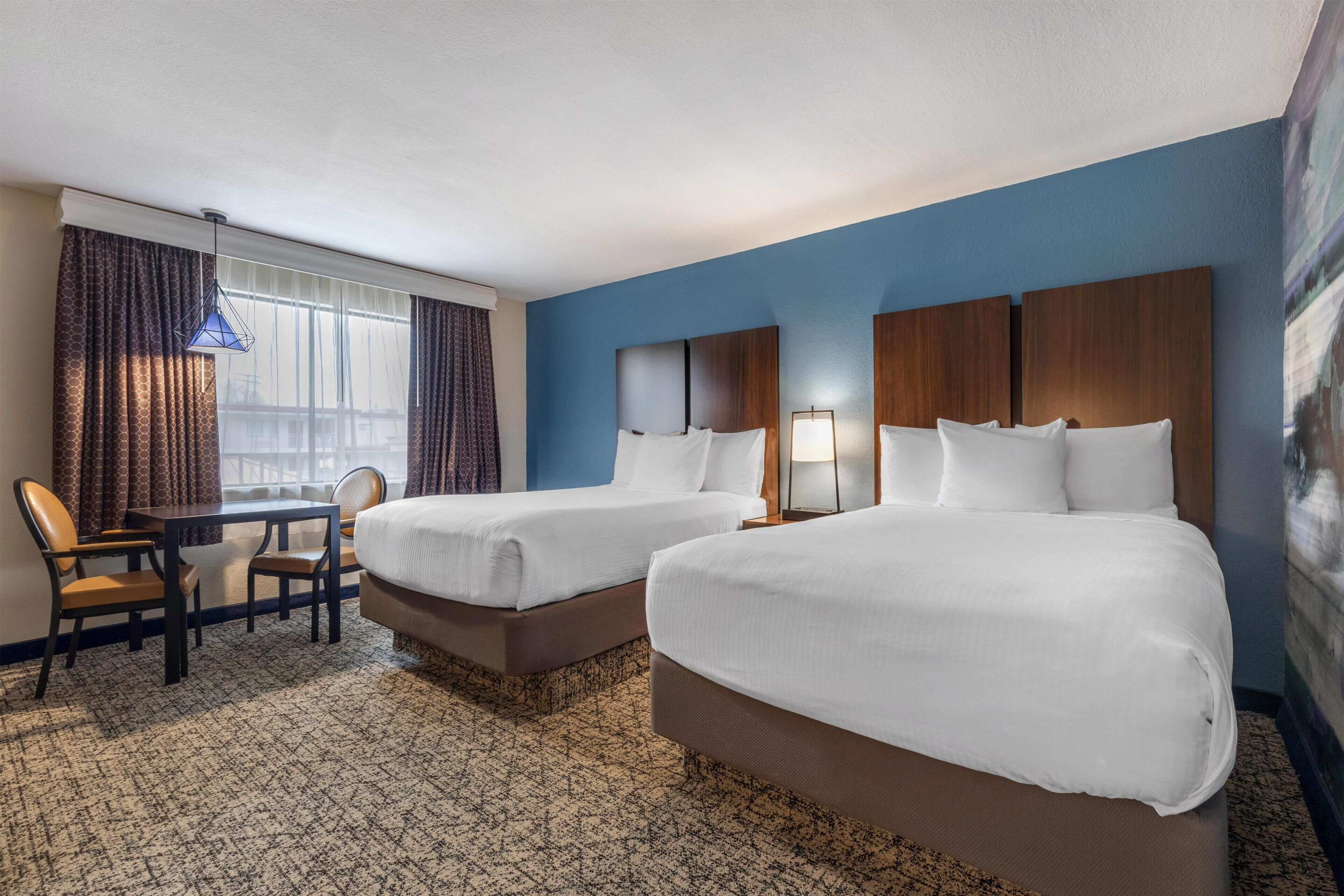 Een hotelkamer met twee bedden, een bureau, stoelen, een lamp en een groot raam met gordijnen.
