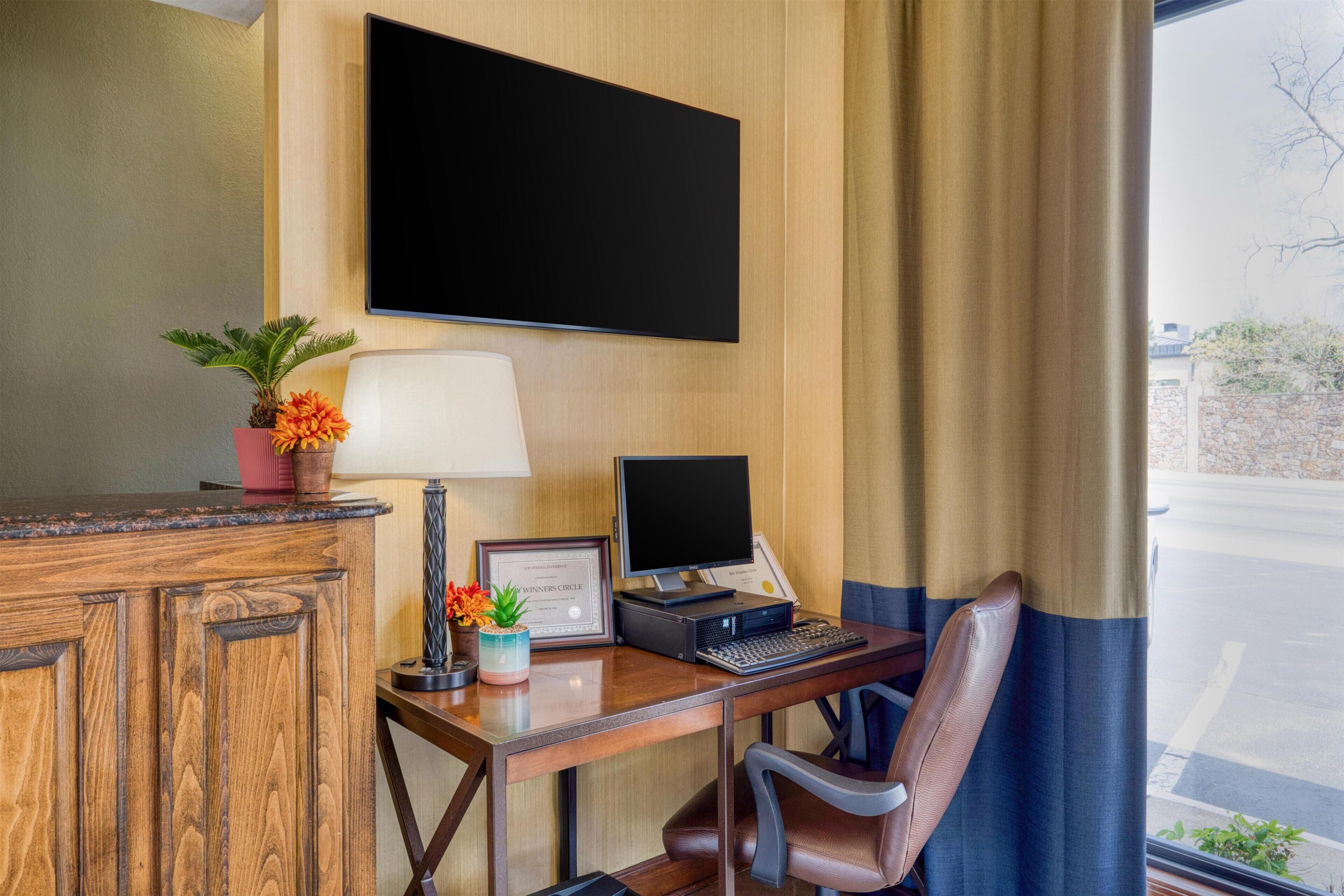 Une chambre d’hôtel avec un bureau en bois, un ordinateur portable, un téléviseur à écran plat, une lampe, une chaise et une fenêtre avec des rideaux.