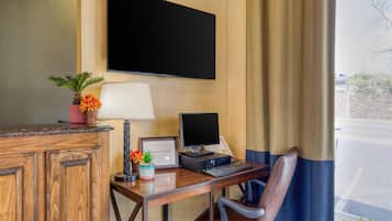 Une chambre d’hôtel avec un bureau en bois, un ordinateur portable, un téléviseur à écran plat, une lampe, une chaise et une fenêtre avec des rideaux.