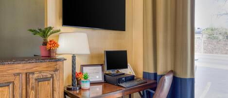 Habitación de hotel con escritorio de madera, laptop, televisor de pantalla plana, lámpara, silla y ventana con cortinas.