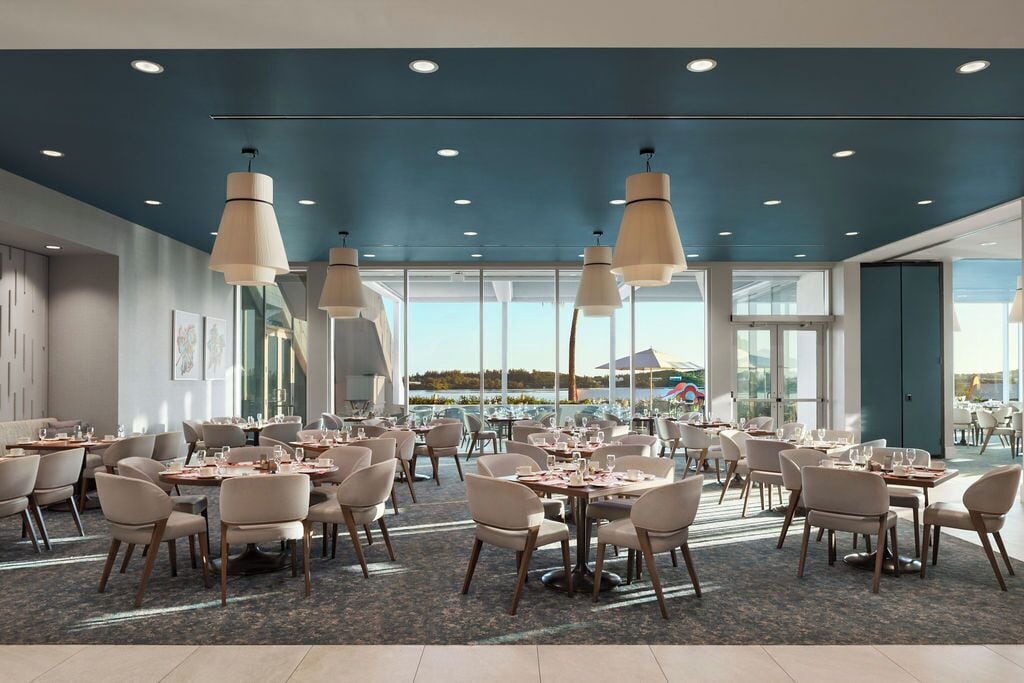 Uma sala de jantar ampla, com várias mesas e cadeiras, luminárias pendentes grandes e vista para o exterior.