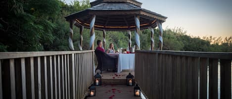 A wooden deck leading to a gazebo with a table set for two, surrounded by lanterns and rose petals.