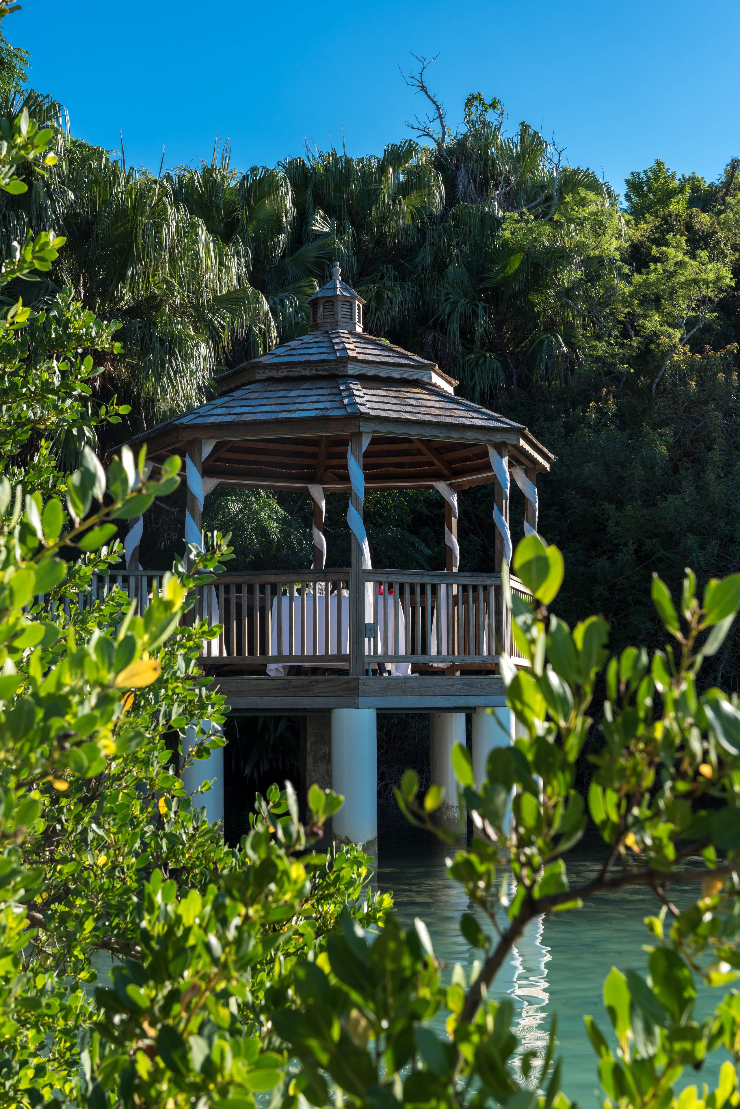 A gazebo with a wooden structure and a balcony, surrounded by lush greenery and a body of water.