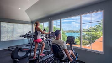 Une salle de sport équipée de tapis de course et d’appareils elliptiques, de grandes fenêtres offrant une vue sur la verdure et un plan d’eau.