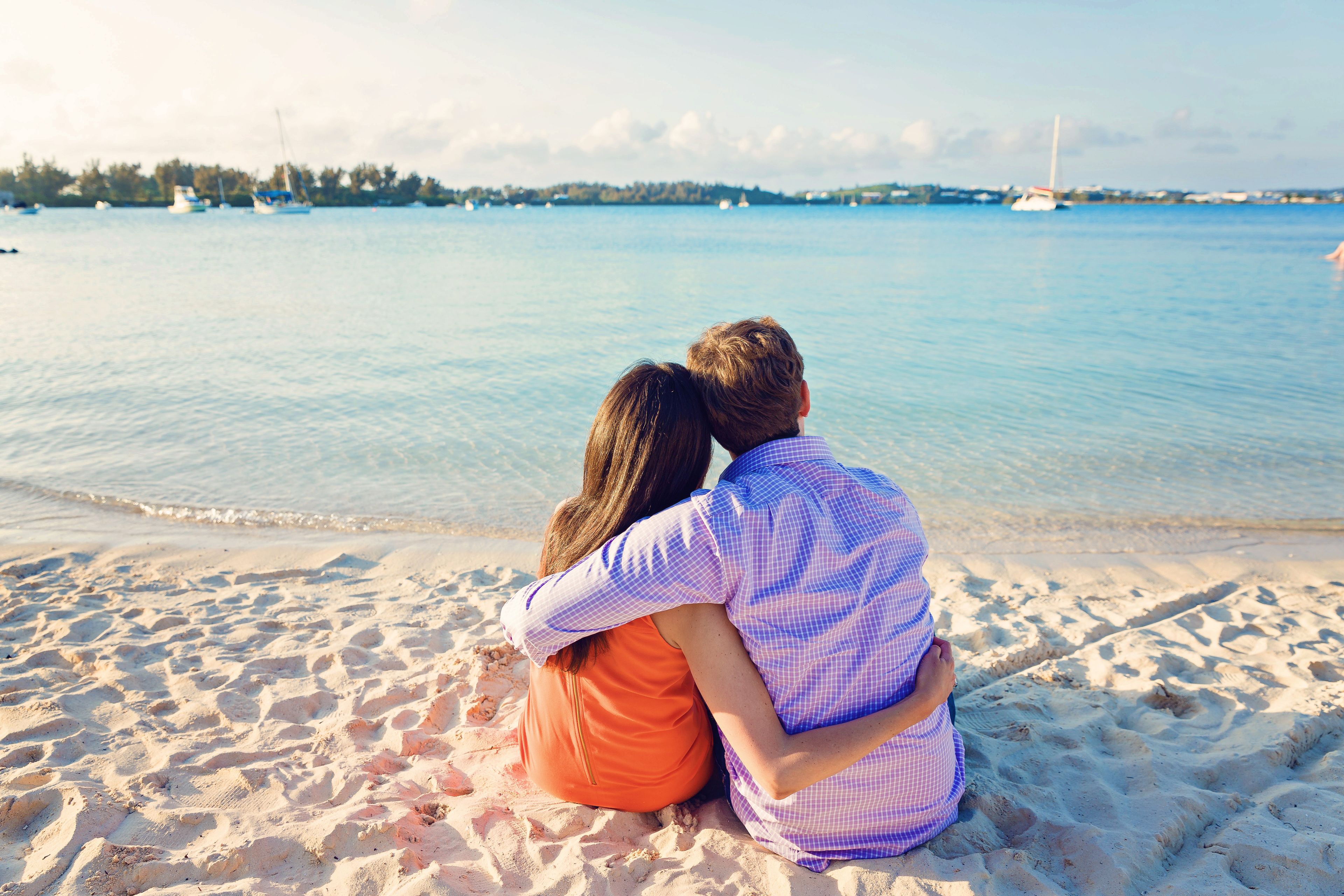 Un couple est assis sur une plage de sable, avec une mer calme et des bateaux en arrière-plan.