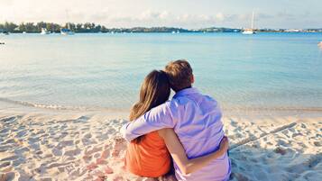 Un couple est assis sur une plage de sable, avec une mer calme et des bateaux en arrière-plan.