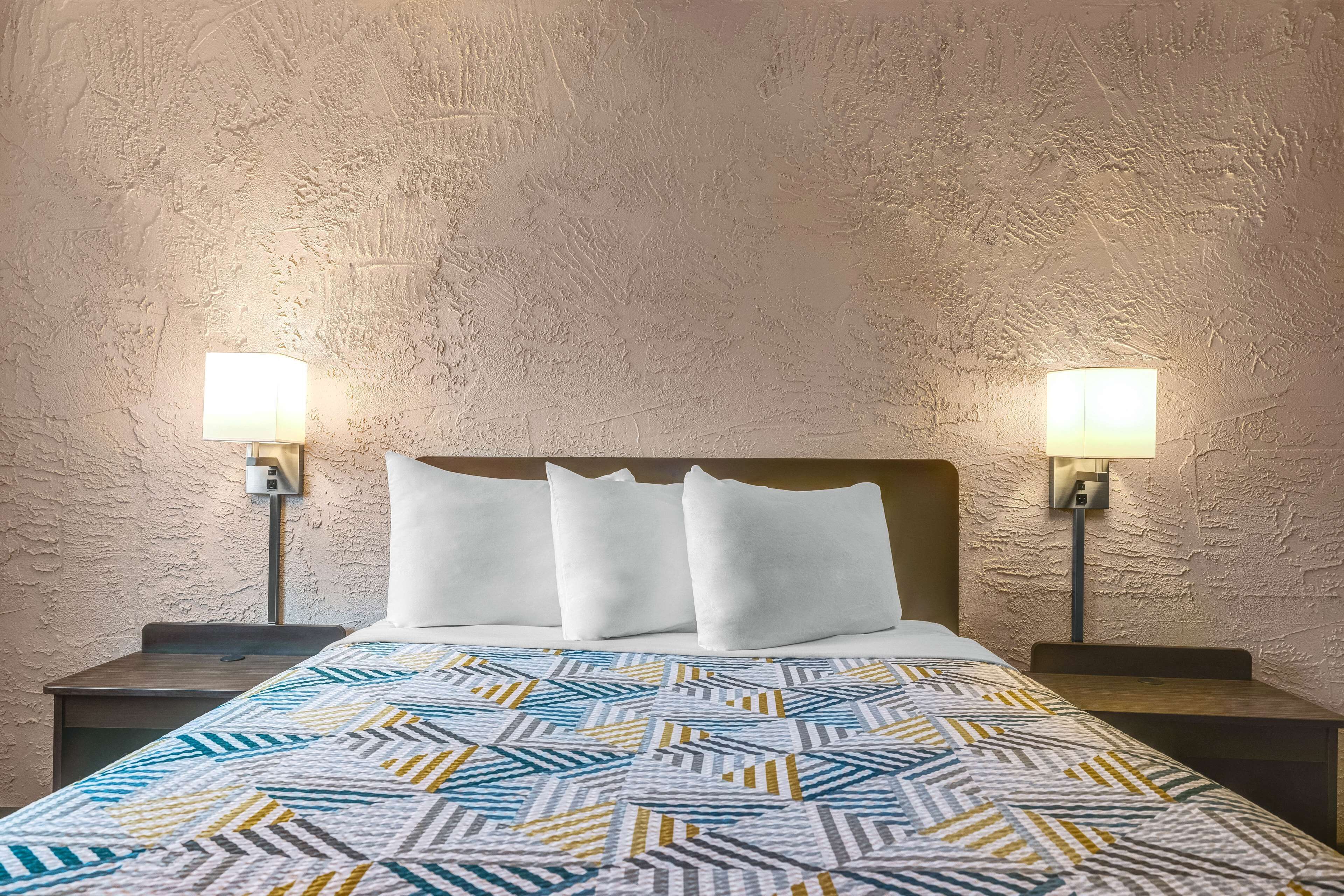 A hotel room with a textured wall, a patterned bedspread, two bedside lamps, and a wooden headboard.