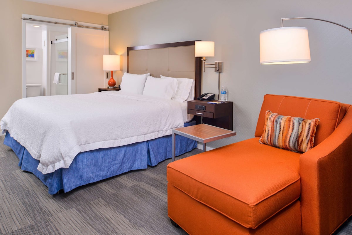 A hotel room with a bed, a nightstand, a desk, an orange armchair, and a lamp.