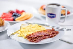 A breakfast plate with scrambled eggs and bacon, a cup of coffee, and a plate of fruit.