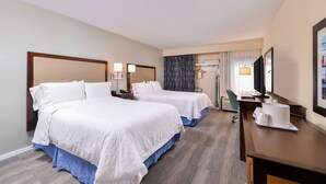 Quarto de hotel com duas camas, uma escrivaninha, uma cadeira, televisão e janela com cortinas.