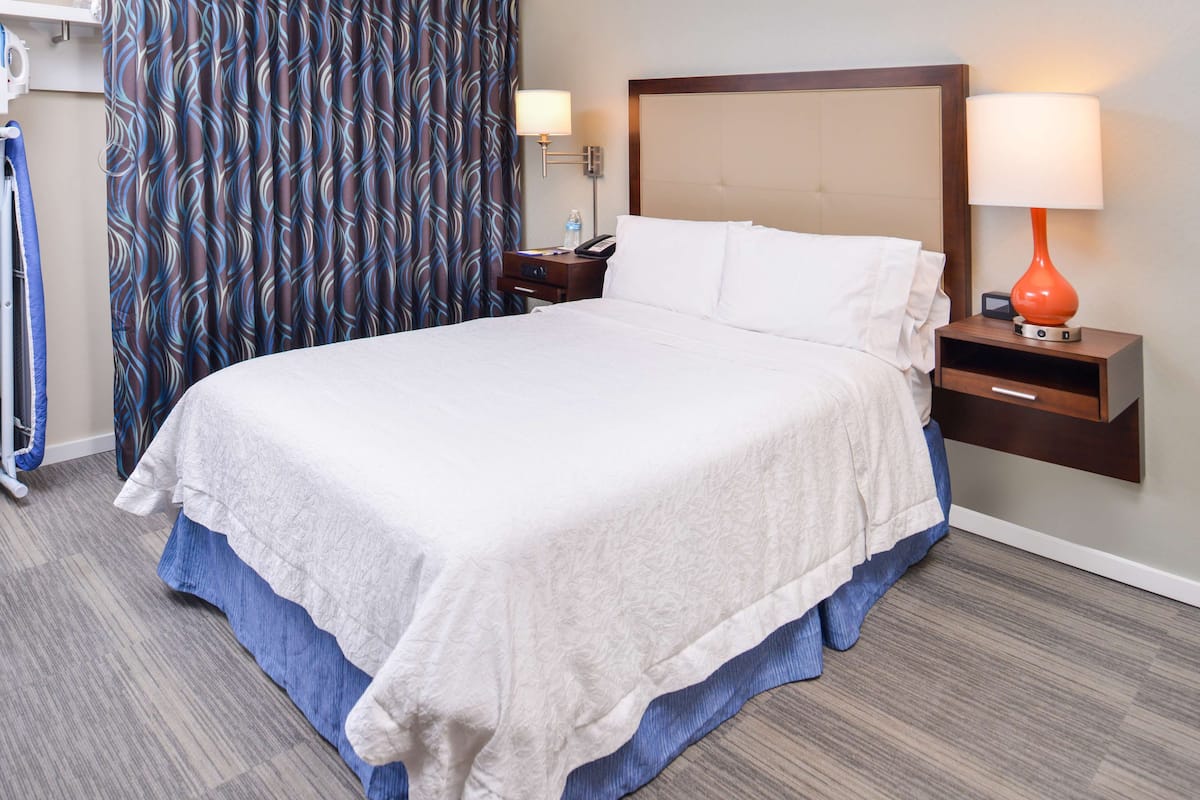 A neatly made bed with white linens, a wooden headboard, a nightstand with a lamp, and a patterned curtain.