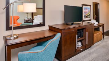 A hotel room with a wooden desk, a blue armchair, a flat-screen TV, a framed picture on the wall, and a suitcase on the floor.
