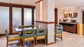 Un comedor con una mesa de madera y sillas tapizadas de verde, una barra con máquinas de café y un cubo de basura.