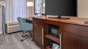 Quarto de hotel com escrivaninha de madeira, cadeira de escritório azul-esverdeada, televisão de tela plana, espelho, luminária e aquecedor.