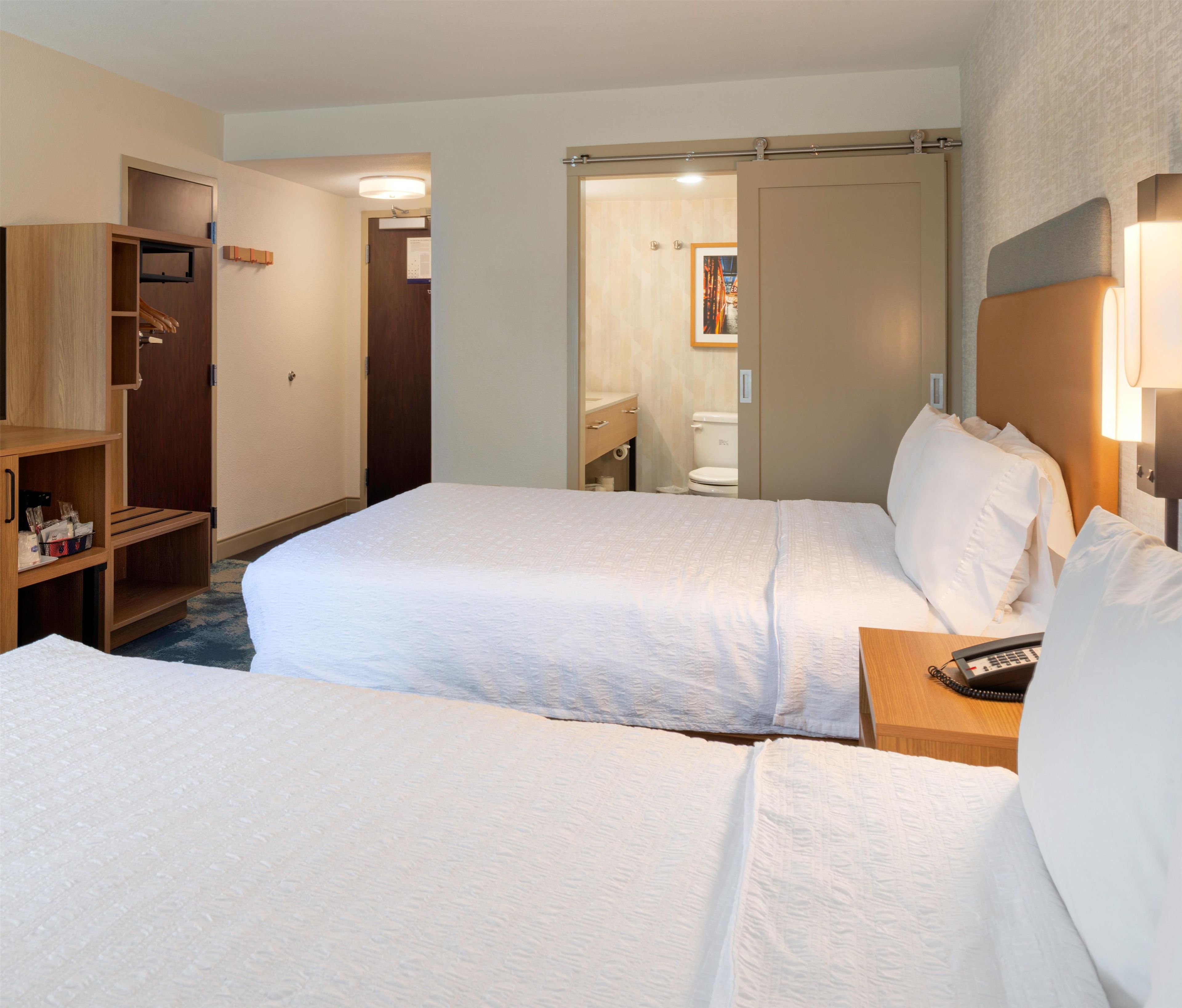 A hotel room with two beds, a wooden wardrobe, a bathroom with a toilet and sink, and a bedside table with a phone.