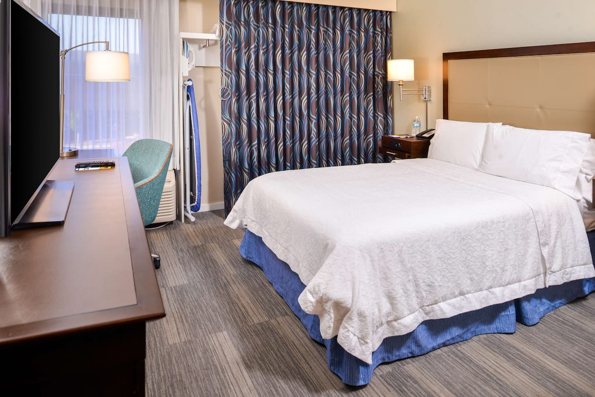 A hotel room with a bed, a desk with a TV, a chair, a nightstand, and a window with curtains.