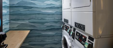 A laundry room with a wall-mounted washing machine and dryer, a wooden bench, and a patterned wall.