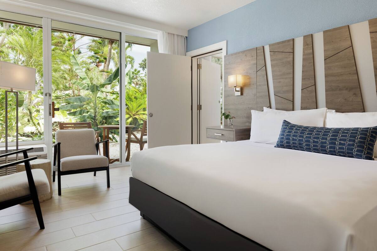Een moderne hotelkamer met een groot bed, een bureau en een stoel. Er is uitzicht op weelderige begroeiing door de open deur.