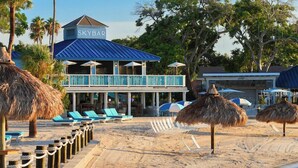 Un hotel frente a la playa con piscina, palmeras y una sombrilla de paja.