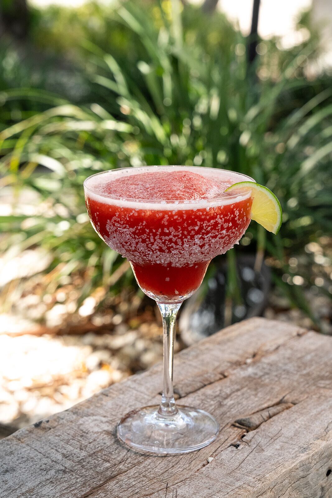 A cocktail in a stemmed glass with a salted rim, garnished with a lime slice, on a wooden surface.