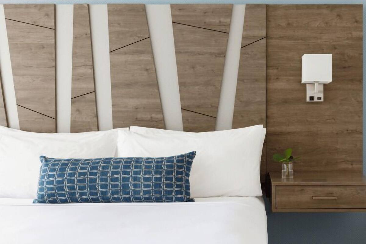Een bed met witte beddengoed, een blauw gestreept kussen en een houten hoofdbord met een lichtschakelaar.