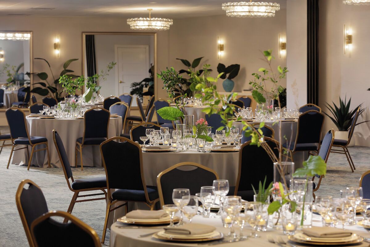 Un salón de banquetes con mesas redondas dispuestas para comer, rodeado de sillas y decorado con plantas y velas.