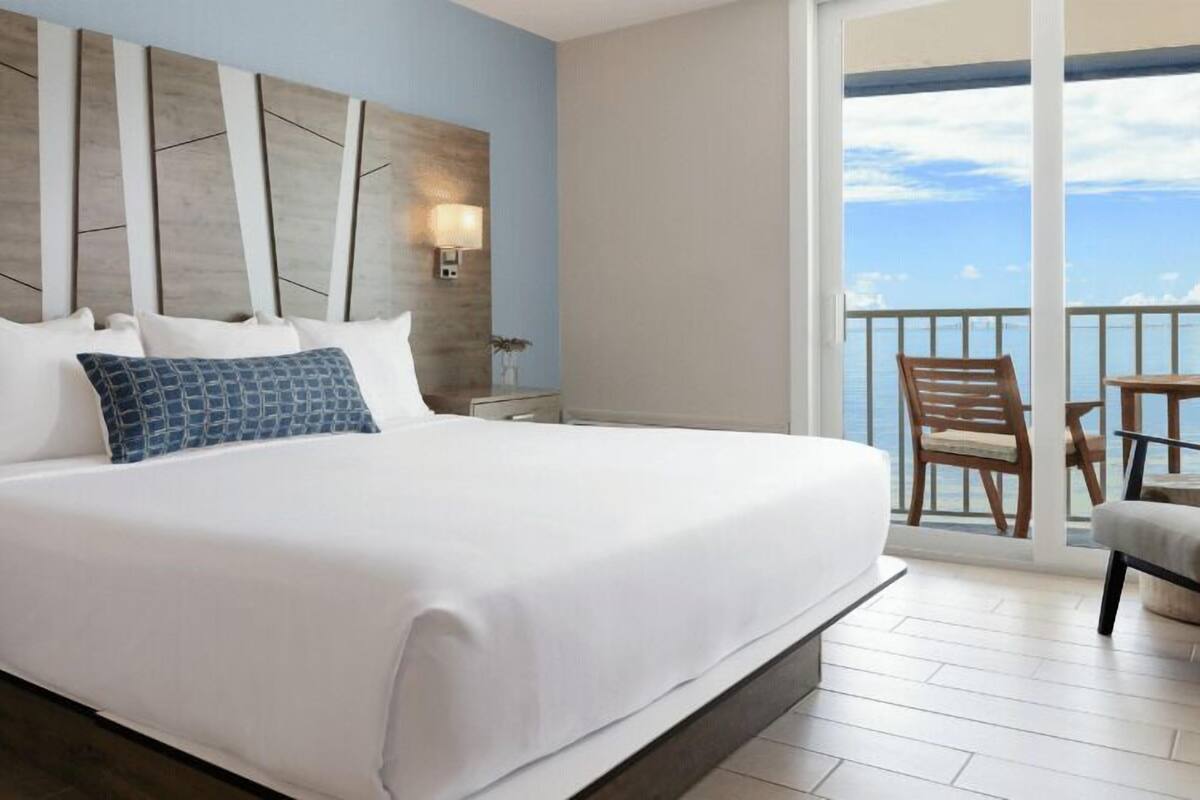 Een groot bed met witte lakens en een blauw gestreept kussen, een houten hoofdbord, een nachtkastje, een wandlamp en uitzicht op de oceaan vanuit een balkon.