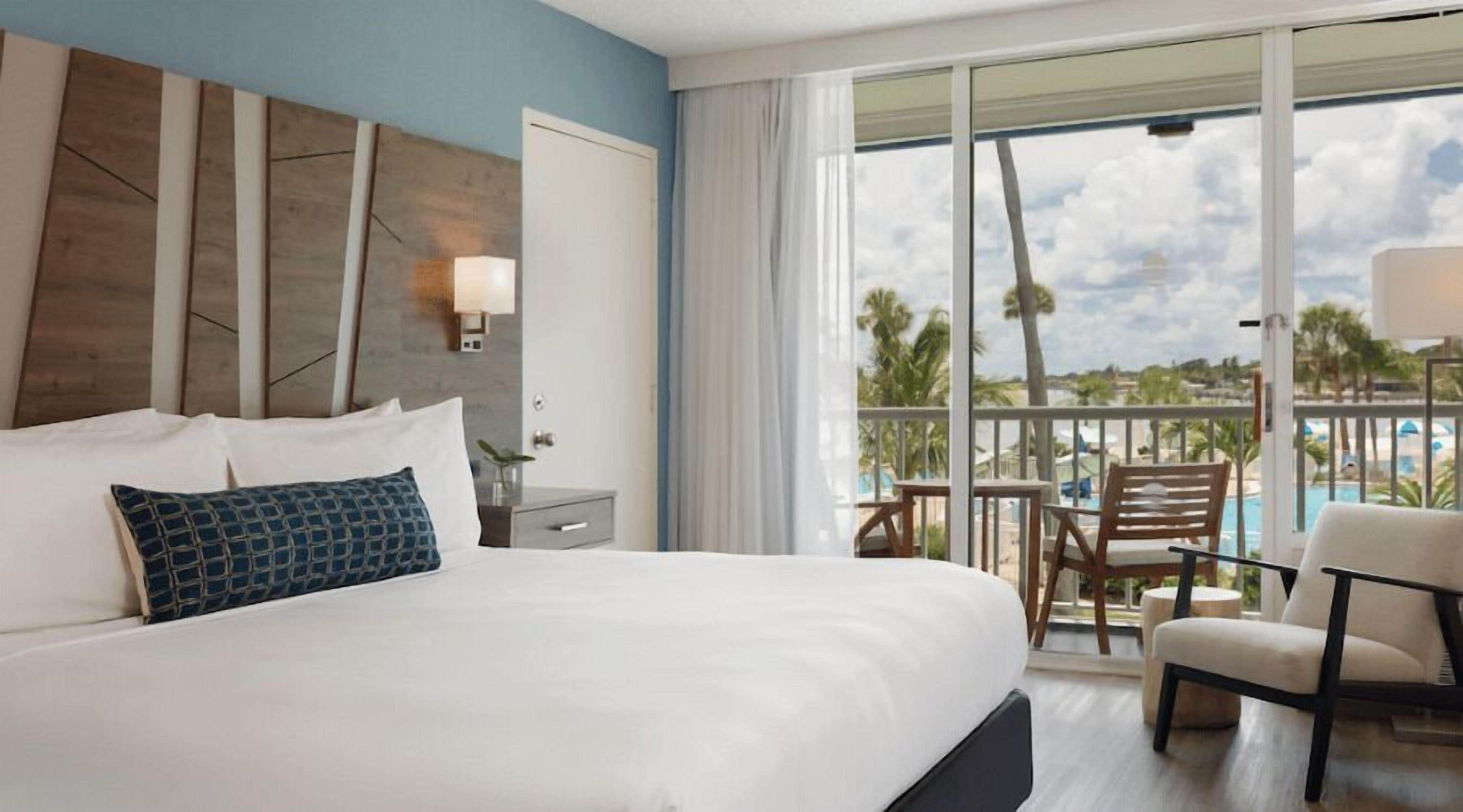 A hotel room with a large bed, a nightstand, a lamp, a view of a pool and palm trees through a balcony.