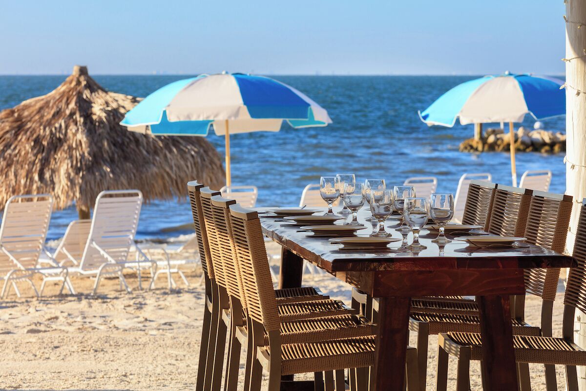 A beachside dining table set with chairs and umbrellas, overlooking the ocean.