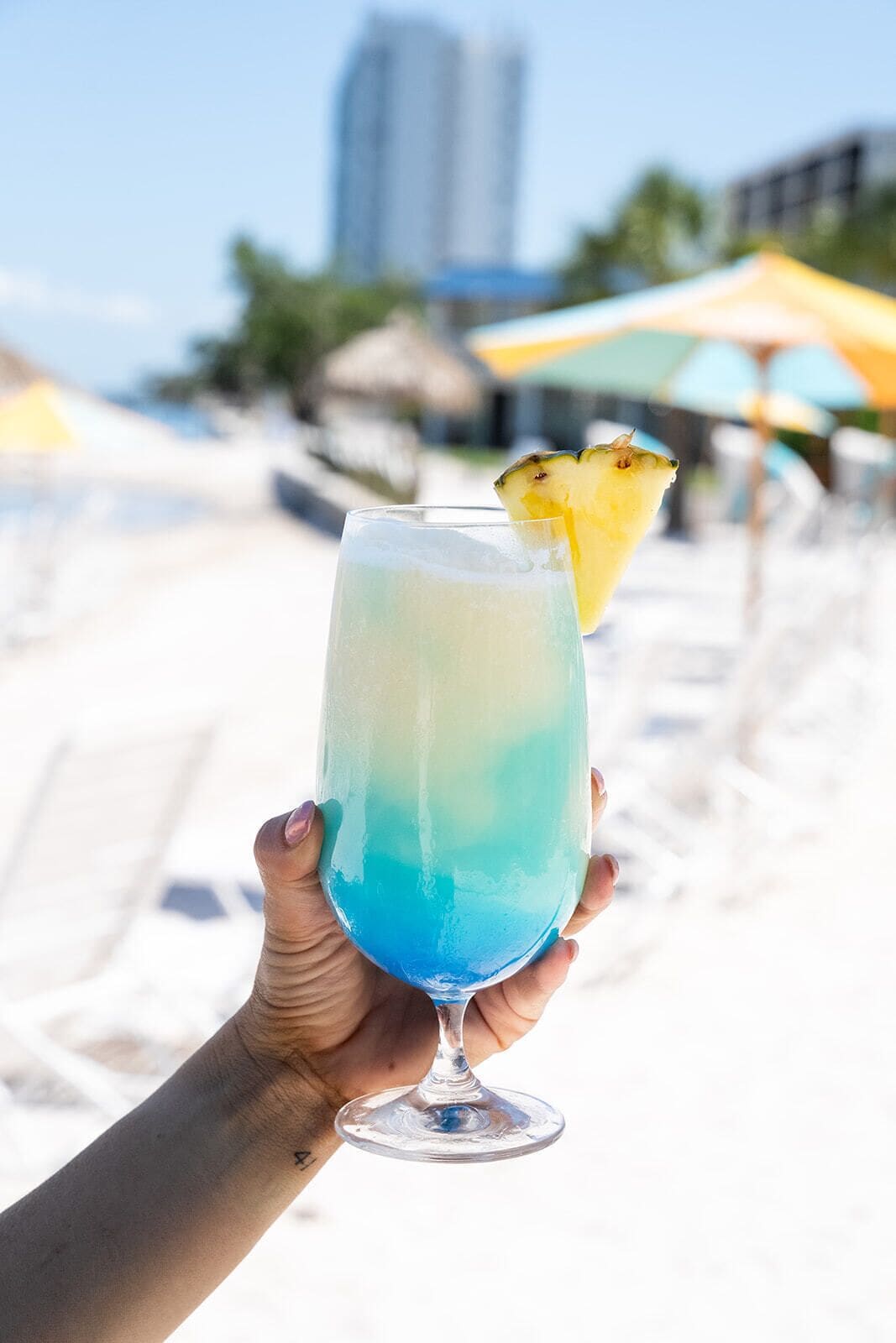 A hand holding a cocktail glass with a gradient color from blue to green, topped with a pineapple slice.